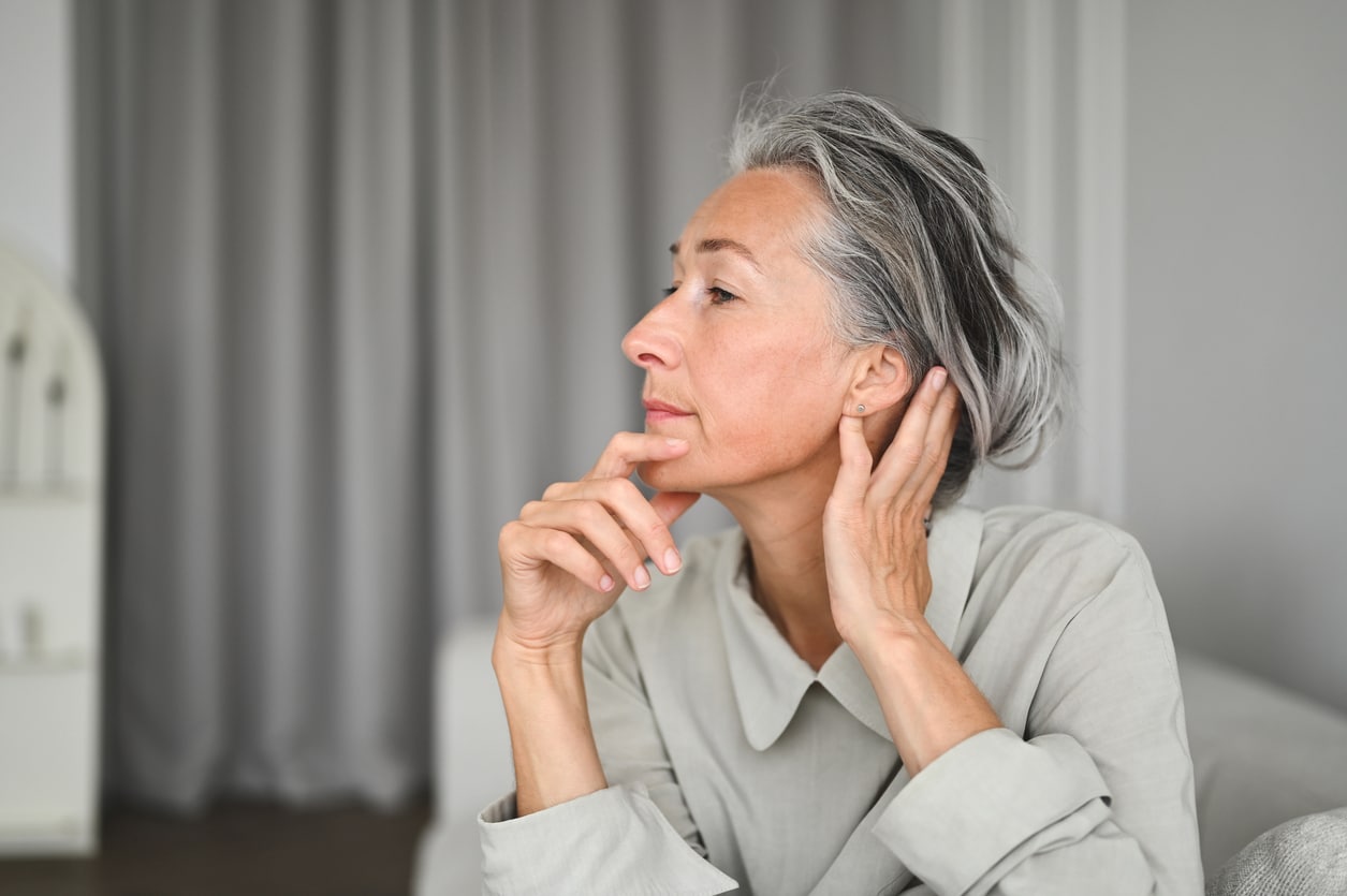 Profile view portrait of elegant senior woman. She is wearing grey with grey hair facing left. The image is a profile and she looks throughtful with her one hand resting on her chin and the other by her ear.