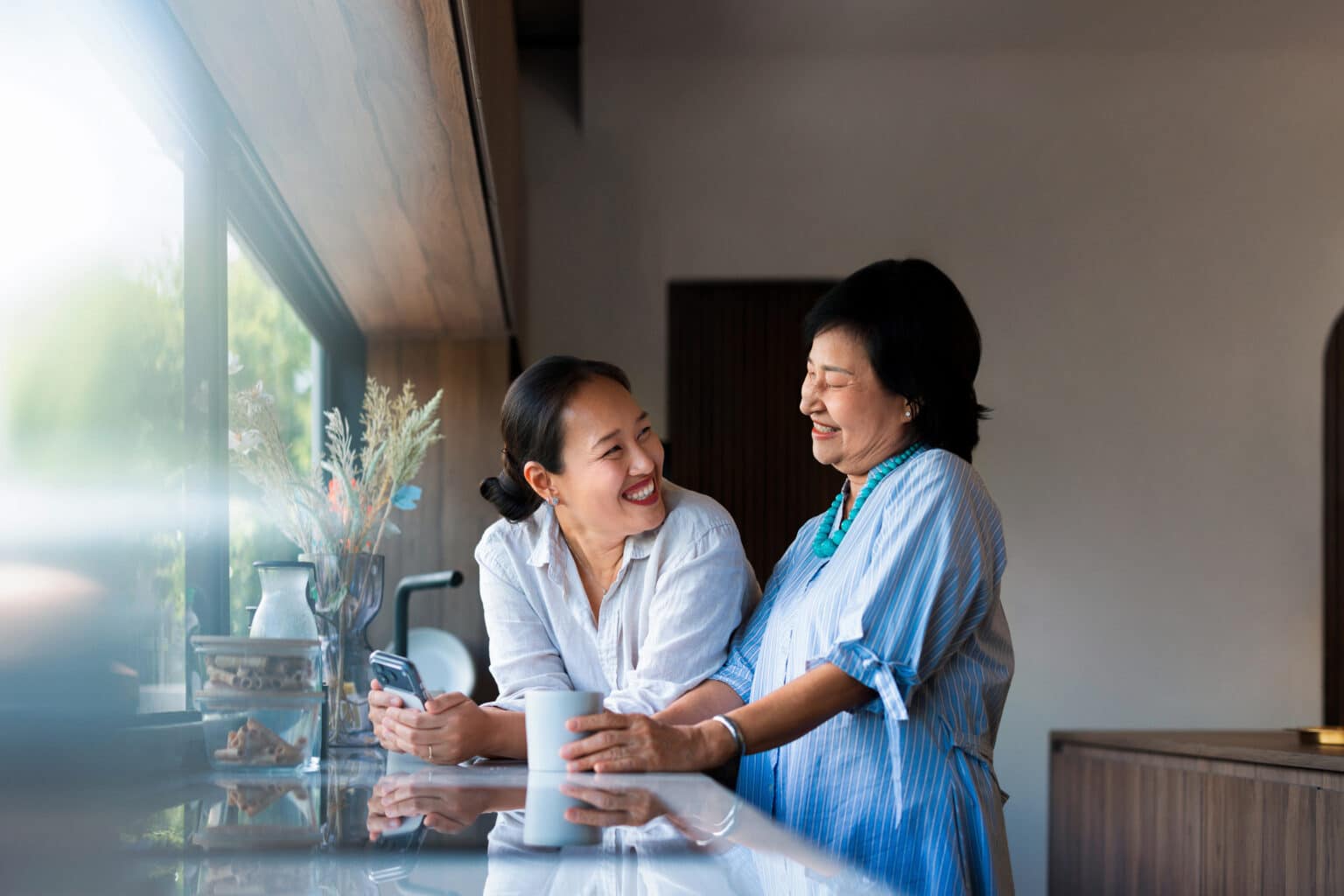 Woman and her adult daughter laughing in the kitchen, enjoying their morning, drinking coffee.