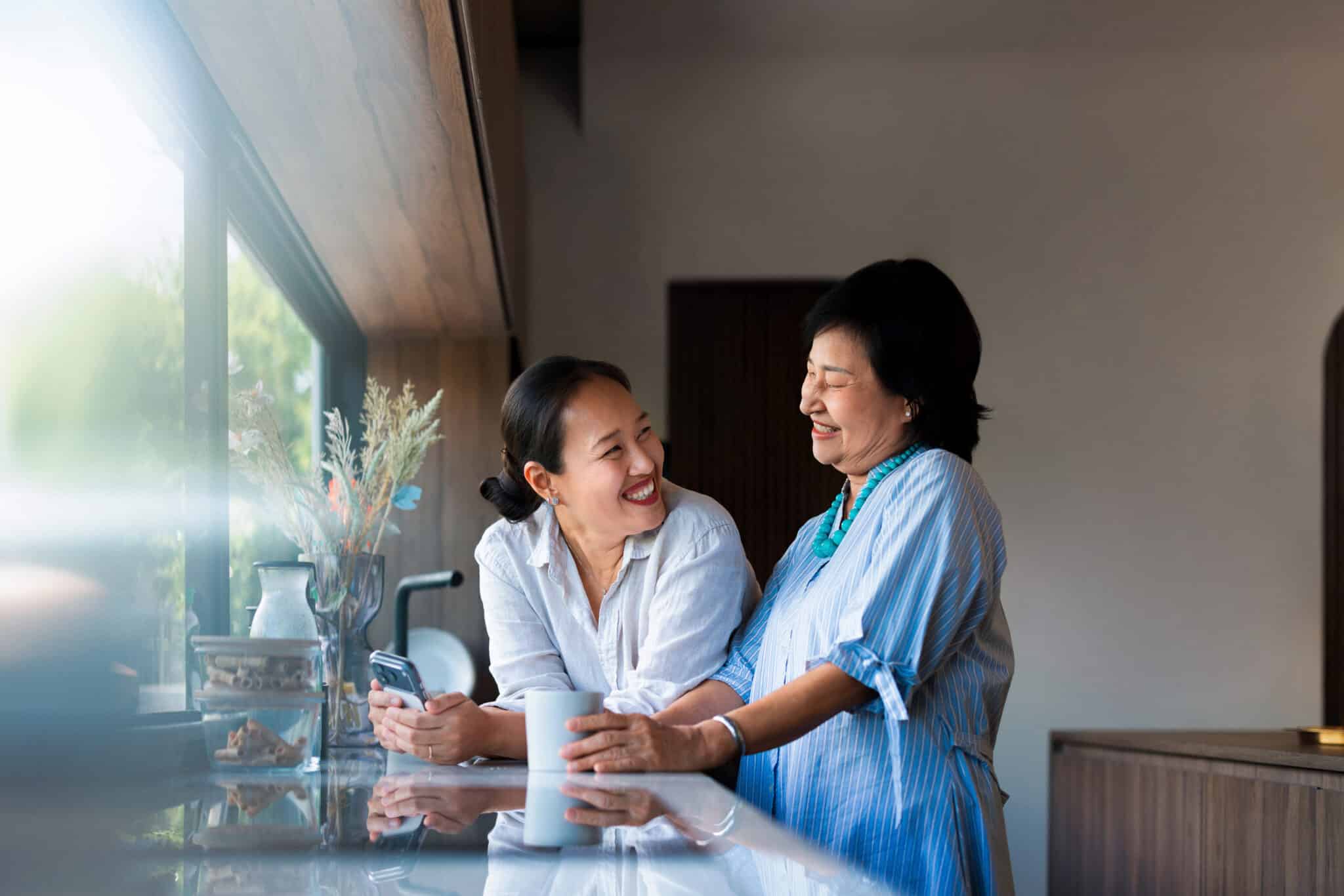 Woman and her adult daughter laughing in the kitchen, enjoying their morning, drinking coffee.